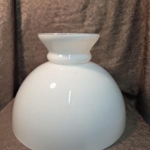 May include: A white glass lamp shade with a smooth, rounded dome shape and a flared, cylindrical neck. The surface of the glass is opaque and features a glossy finish that reflects light, creating a clean and simple appearance for lighting fixtures.