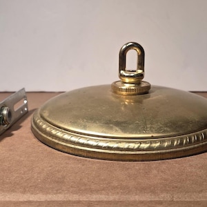 May include: A circular brass ceiling canopy with a central loop and a decorative rope-patterned edge. Beside the canopy is a silver-toned metal mounting bracket with a threaded bolt and nut, designed for securing the fixture to a ceiling junction box.