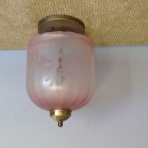 May include: A vintage ceiling light fixture with a frosted pink glass shade. The shade has a floral design and is accented with brass hardware. The light is mounted on a beige surface and a white wall.