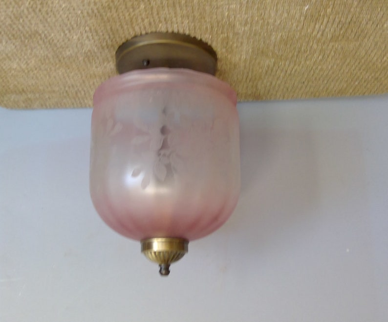 May include: A vintage ceiling light fixture with a frosted pink glass shade. The shade has a floral design and is accented with brass hardware. The light is mounted on a beige surface and a white wall.