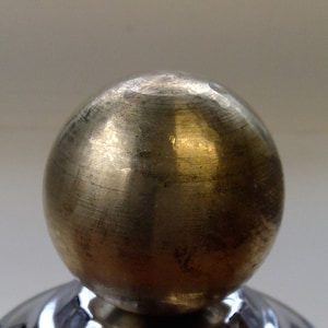 May include: A close-up of a brass sphere with a textured surface. The sphere is part of a decorative object, possibly a door knob or furniture accent. The brass has a warm, golden hue.