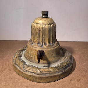 May include: A vintage brass lamp base with a decorative design. The base has a round, ornate bottom with a leaf pattern, a fluted midsection, and a small, threaded top. A small, curved metal piece is attached to the side.