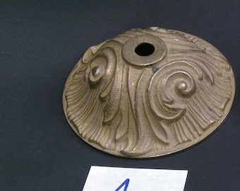 May include: A detailed, antique brass-colored metal decorative piece with an ornate, swirling leaf design. The circular object has a central hole and is likely a decorative element for furniture or lighting. A white paper with the letter "A" is in the foreground.