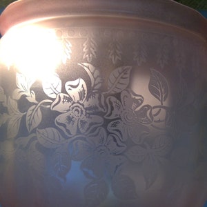 May include: A frosted glass vase with etched floral and leaf designs. The vase has a wide, rounded body and a slightly flared rim. The floral pattern includes large, detailed flowers and delicate leaves. The vase is lit from within, creating a soft glow.