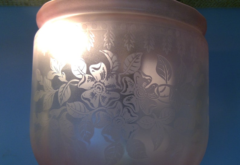 May include: A frosted glass vase with etched floral and leaf designs. The vase has a wide, rounded body and a slightly flared rim. The floral pattern includes large, detailed flowers and delicate leaves. The vase is lit from within, creating a soft glow.