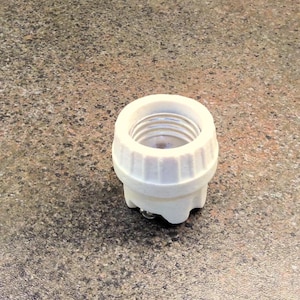 May include: A white ceramic light bulb socket with a ribbed exterior and a metal interior. The socket is designed to hold a light bulb and has a small base.