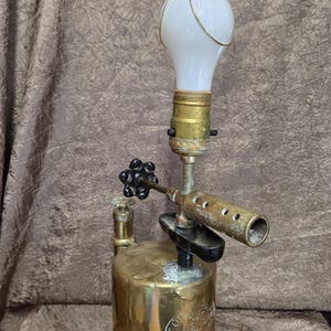 May include: A unique lamp crafted from a vintage brass blowtorch, featuring a white bulb and intricate metalwork. The lamp has a cylindrical base and a long, curved nozzle, showcasing a blend of industrial design and functionality.