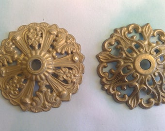May include: Two antique brass-colored ceiling medallions with intricate floral and scrollwork designs. Each medallion has a central hole and radiating patterns, suggesting a vintage or decorative lighting fixture component.