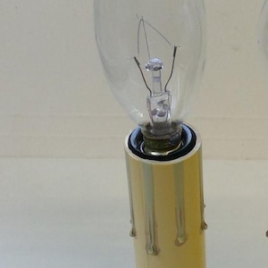 May include: A clear, flame-shaped light bulb with a visible internal filament, mounted on a cylindrical, pale yellow plastic base designed to resemble a dripping wax candle, featuring a metallic socket connection at the top of the base.