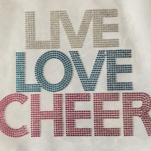 May include: White fabric with the words "LIVE LOVE CHEER" spelled out in rhinestones. The "LIVE" is in white rhinestones, the "LOVE" is in blue rhinestones, and the "CHEER" is in pink rhinestones.