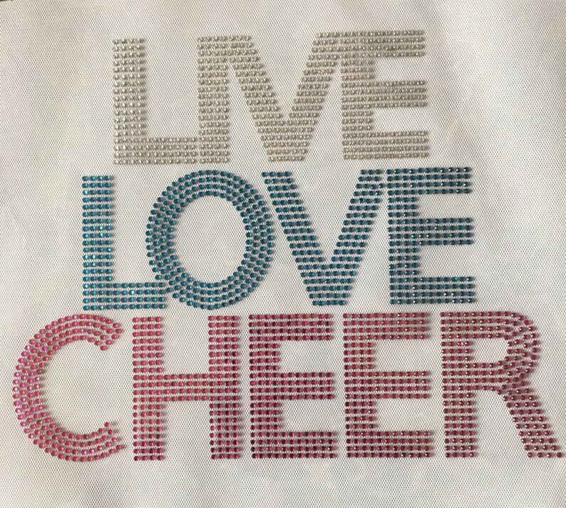 May include: White fabric with the words "LIVE LOVE CHEER" spelled out in rhinestones. The "LIVE" is in white rhinestones, the "LOVE" is in blue rhinestones, and the "CHEER" is in pink rhinestones.