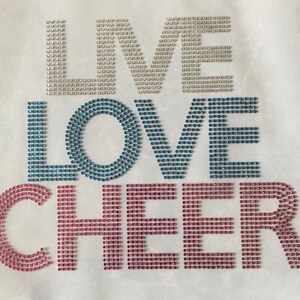 May include: White fabric with the words "LIVE LOVE CHEER" spelled out in rhinestones. The "LIVE" is in silver, "LOVE" is in blue and teal, and "CHEER" is in pink.