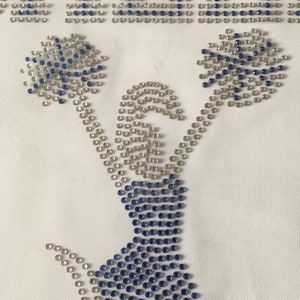 May include: A white fabric with a blue and silver rhinestone design of a cheerleader holding pom-poms. The word "CHEER" is spelled out in rhinestones at the top of the design.