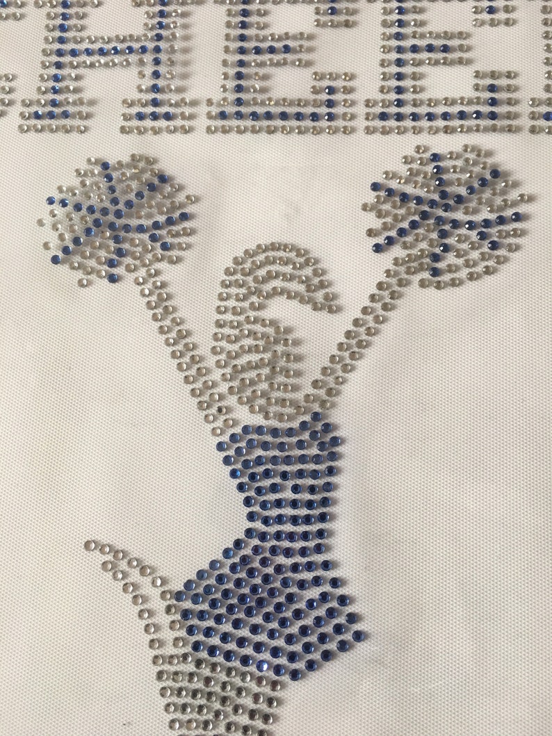 May include: A white fabric with a blue and silver rhinestone design of a cheerleader holding pom-poms. The word "CHEER" is spelled out in rhinestones at the top of the design.
