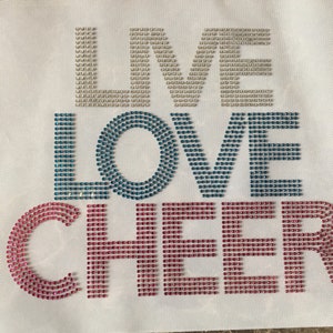 May include: A white fabric background with the words "I LOVE CHEER" spelled out in rhinestones. The letters "I" and "LOVE" are in light blue and teal rhinestones, and the word "CHEER" is in pink rhinestones.