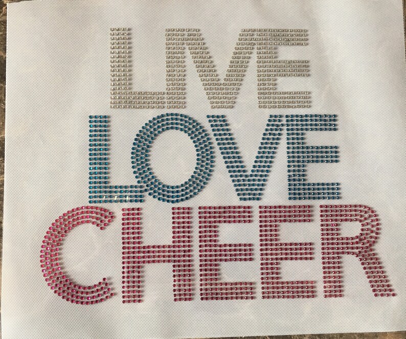 May include: A white fabric background with the words "I LOVE CHEER" spelled out in rhinestones. The letters "I" and "LOVE" are in light blue and teal rhinestones, and the word "CHEER" is in pink rhinestones.