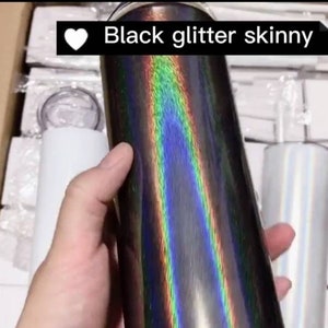 May include: A black glitter skinny tumbler with an iridescent finish. The tumbler is being held by a hand and is surrounded by other tumblers.