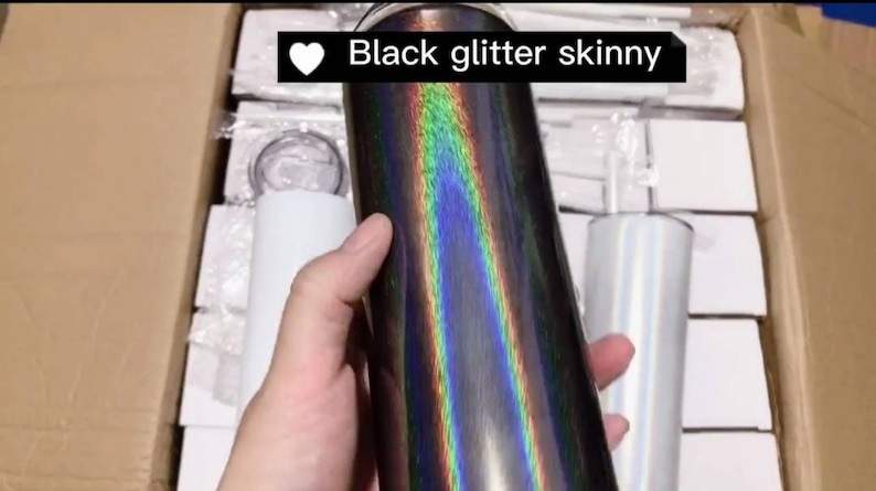 May include: A black glitter skinny tumbler with an iridescent finish. The tumbler is being held by a hand and is surrounded by other tumblers.