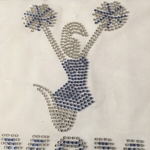 May include: A white fabric with a blue and silver rhinestone design of a cheerleader holding pom-poms. The word "MOM" is spelled out in rhinestones below the cheerleader.