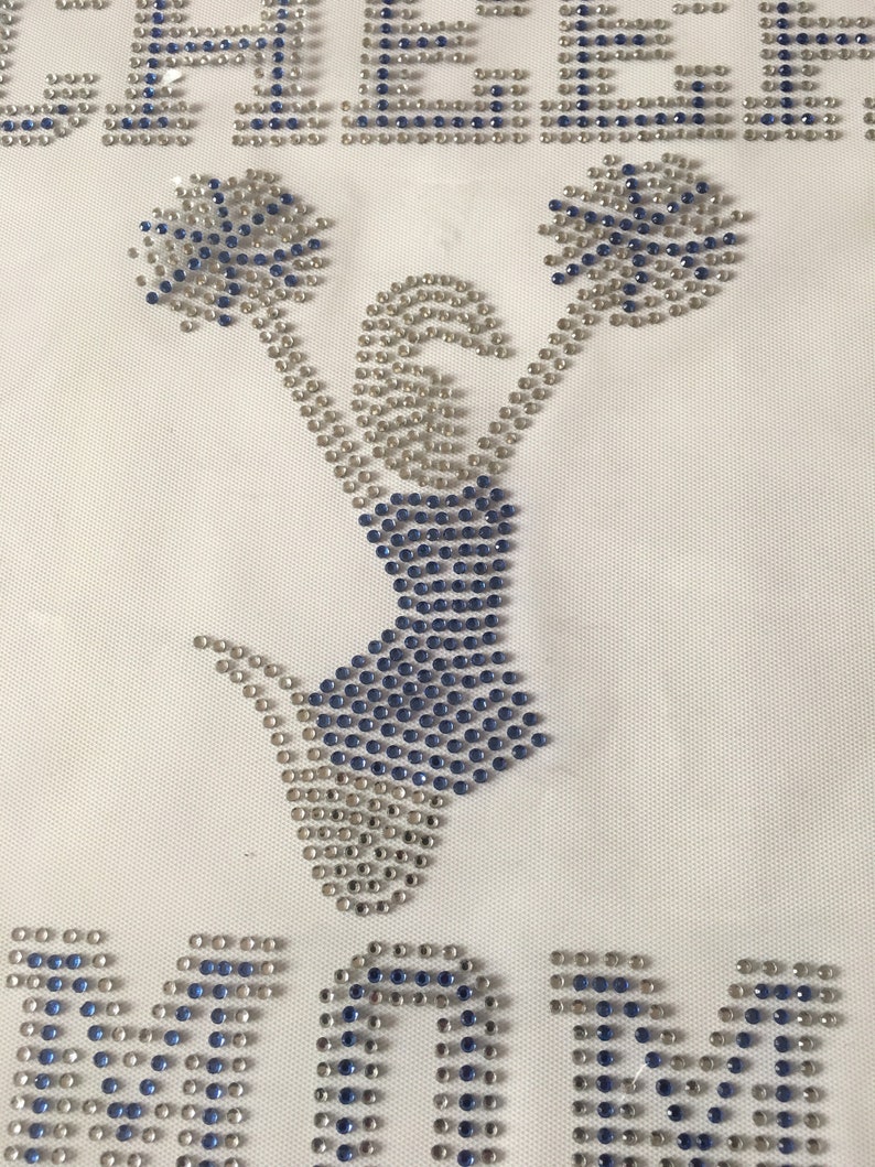 May include: A white fabric with a blue and silver rhinestone design of a cheerleader holding pom-poms. The word "MOM" is spelled out in rhinestones below the cheerleader.