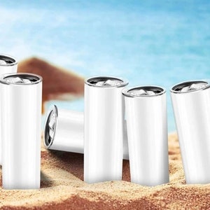 Puede incluir: Seis vasos de acero inoxidable blanco con tapas plateadas, dispuestos sobre un fondo de playa de arena.