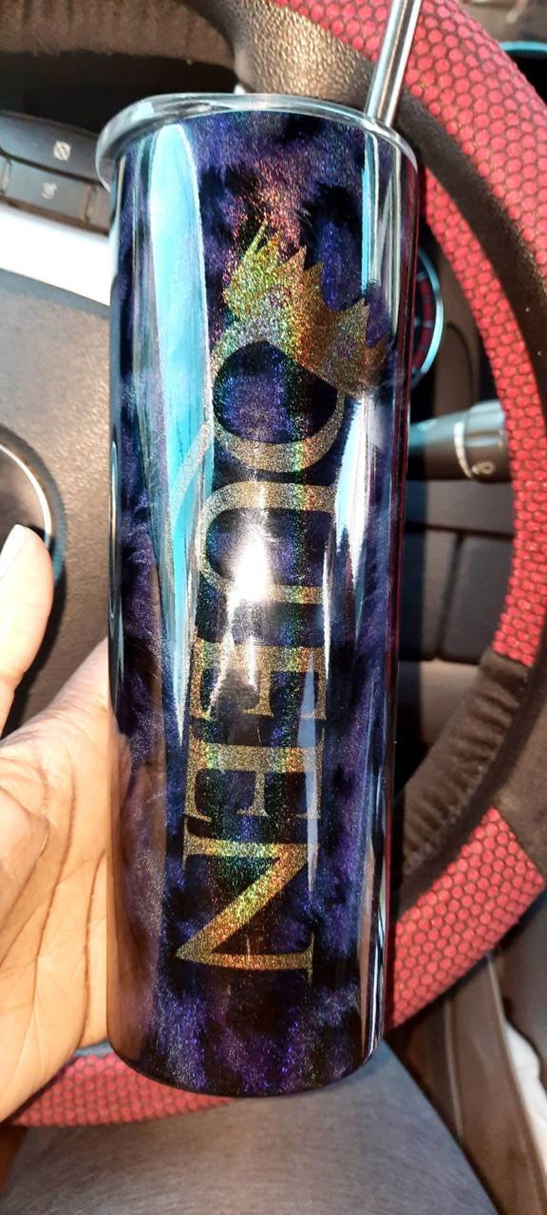 May include: A black and blue tumbler with a gold glitter design and the word "QUEEN" written in gold glitter. The tumbler has a straw and a lid.