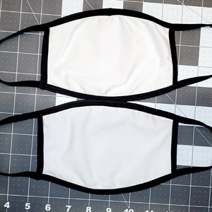 May include: Two white face masks with black elastic ear loops and black trim around the edges. The masks are lying flat on a gray surface.