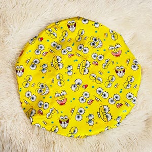 May include: A yellow satin bonnet with a repeating pattern of cartoon character faces. The design features blue eyes, red accents, and white teeth on a bright yellow background. The bonnet is circular and made of a smooth, shiny fabric.