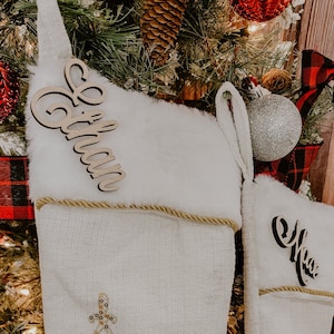 May include: Two white Christmas stockings with gold trim and sequined snowflakes. The stockings have wooden name tags that say "Ethan" and "Max".
