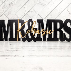 May include: Black wooden "Mr. & Mrs." sign with a gold cursive "Kobasic" in the center. The sign is on a white wooden surface.