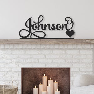May include: Black metal sign with the name "Johnson" in a cursive font with a heart at the end. The sign is mounted above a white brick fireplace with candles inside.