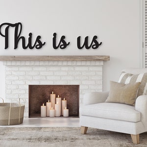 May include: Black wooden sign with the words "This is us" in a cursive font, mounted above a white brick fireplace with a lit fireplace and candles. A white armchair with striped pillows sits in front of the fireplace.