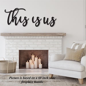 Op de afbeelding: Zwarte metalen wanddecoratie met de woorden "This is Us" in een cursief schrift. De decoratie is geplaatst boven een witte bakstenen open haard met een houten schouw. De open haard heeft kaarsen erin.