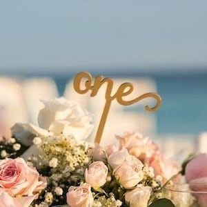 May include: A gold table number with the word "one" in cursive script, placed in a gold vase filled with pink and white roses and baby's breath.