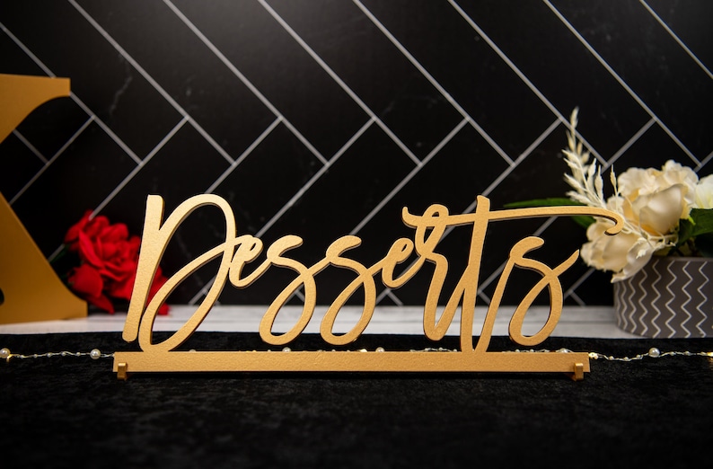 May include: A gold-coloured wooden sign with the word "Desserts" written in a cursive font. The sign is on a black surface with a white background.