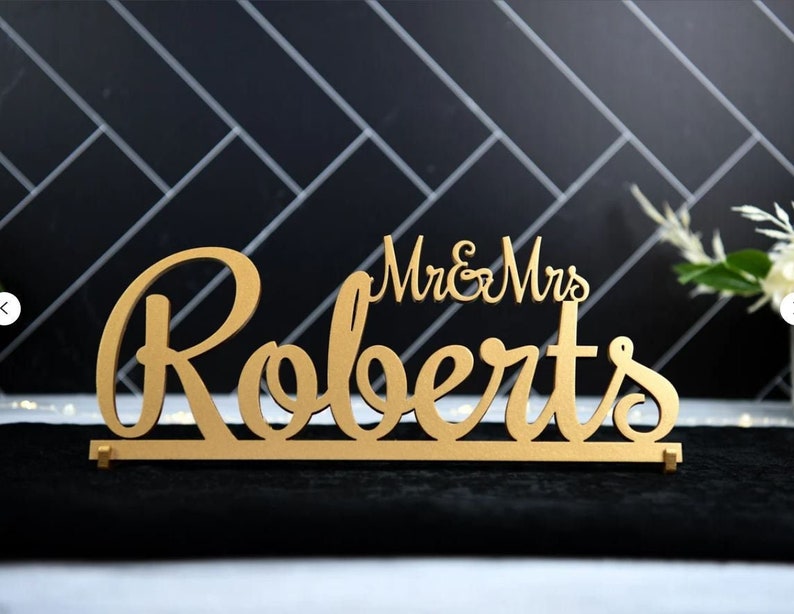 May include: Gold-colored wooden sign with the text "Mr. & Mrs. Roberts" in a cursive font.