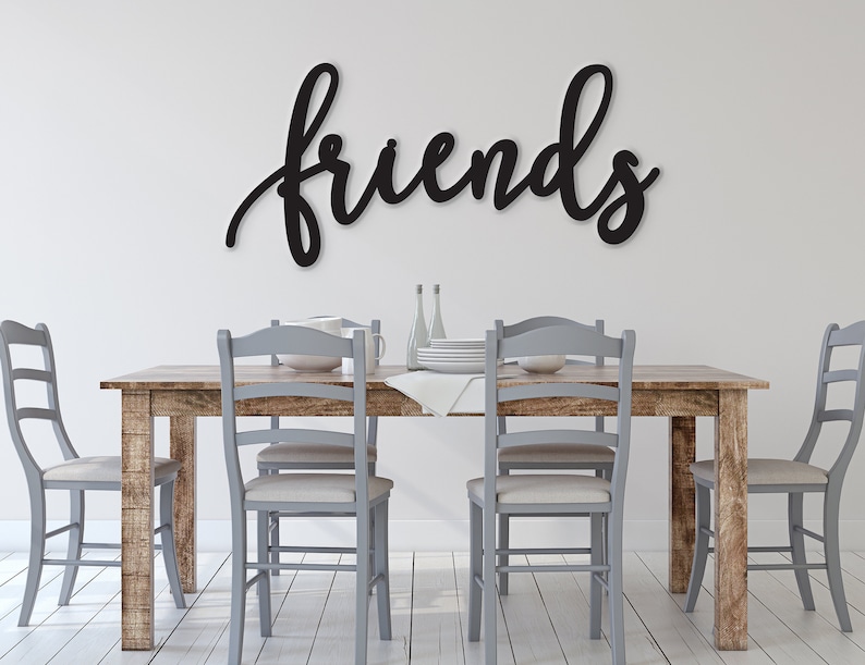May include: A wooden dining table with four grey chairs and a black wooden sign that says "friends" hanging on the wall above the table.