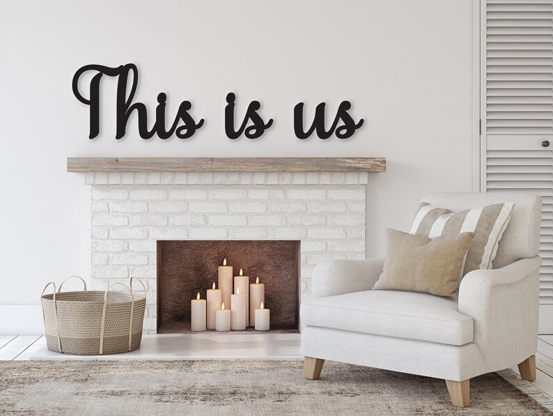 May include: Black wooden sign with the words "This is us" in a cursive font, mounted above a white brick fireplace with a lit fireplace and candles. A white armchair with striped pillows sits in front of the fireplace.
