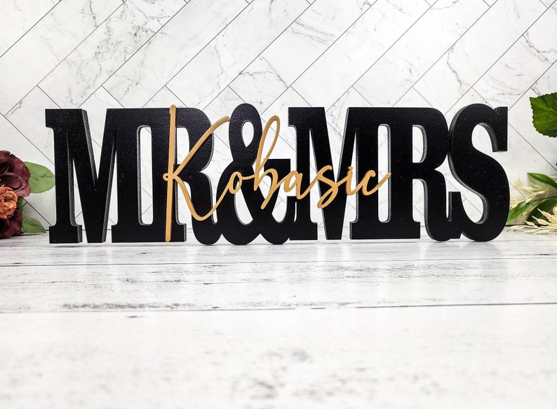 May include: Black wooden "Mr. & Mrs." sign with a gold cursive name "Kobasic" in the center.