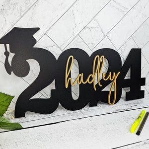 May include: Black wooden 2024 graduation sign with a gold graduation cap and the name "hadley" in gold lettering.