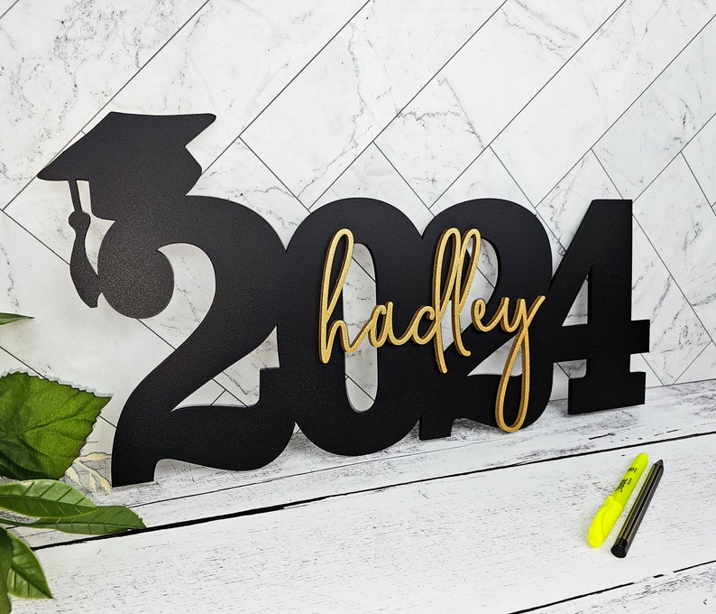 May include: Black wooden 2024 graduation sign with a gold graduation cap and the name "hadley" in gold lettering.