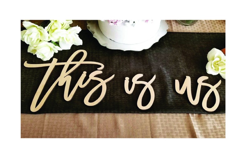 May include: Wooden sign with the words "this is us" in a cursive font. The sign is on a black tablecloth with white flowers on either side.