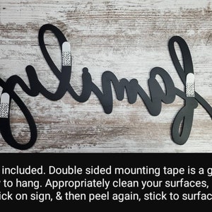 May include: Black metal wall decor with the word "julimah" written in a cursive script. The decor is attached to a white wood background with double-sided mounting tape.