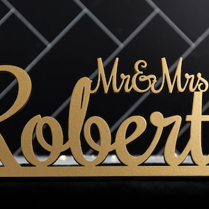 May include: Gold wooden sign with the text "Mr. & Mrs. Roberts" in a cursive font.