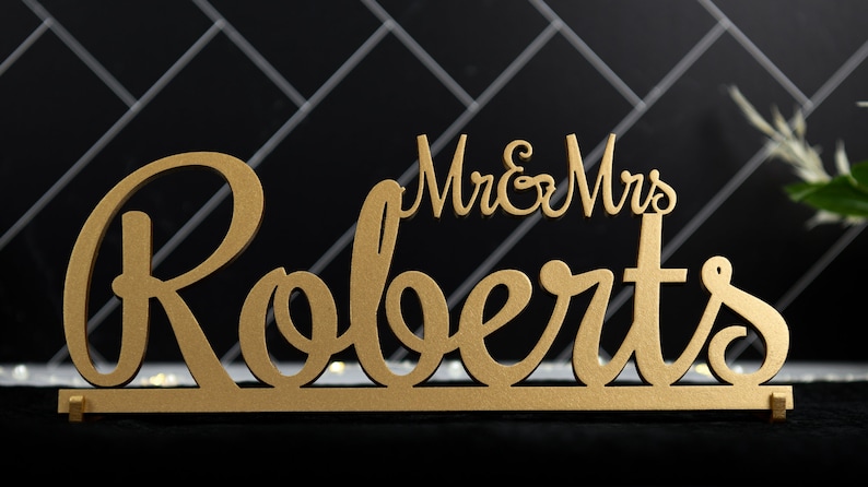 May include: Gold wooden sign with the text "Mr. & Mrs. Roberts" in a cursive font.
