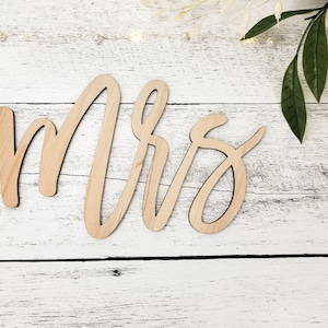 May include: Wooden sign with the word "Mrs." written in cursive script. The sign is light brown and has a natural wood finish.
