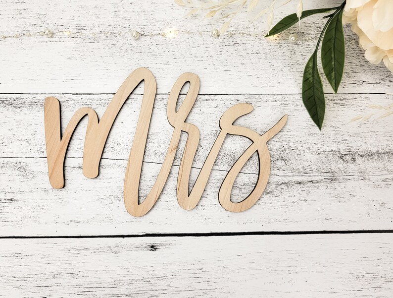 May include: Wooden sign with the word "Mrs." written in cursive script. The sign is light brown and has a natural wood finish.
