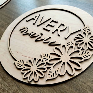 May include: A wooden round sign with the name "Avery Marie" written in a decorative font. The sign is decorated with a floral design around the edge.