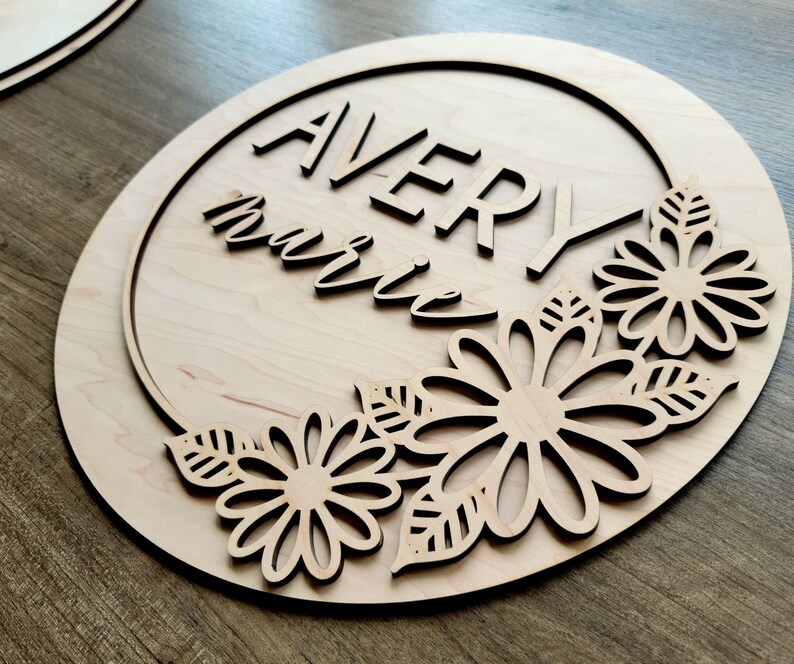 May include: A wooden round sign with the name "Avery Marie" written in a decorative font. The sign is decorated with a floral design around the edge.