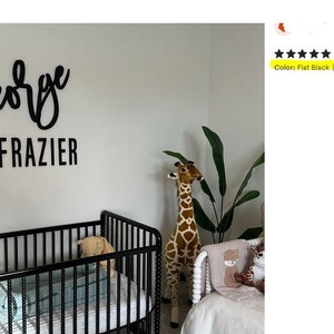May include: Black wooden sign with the name "George Frazier" in cursive script. The sign is mounted on a white wall in a nursery. A stuffed giraffe and a crib are visible in the background.